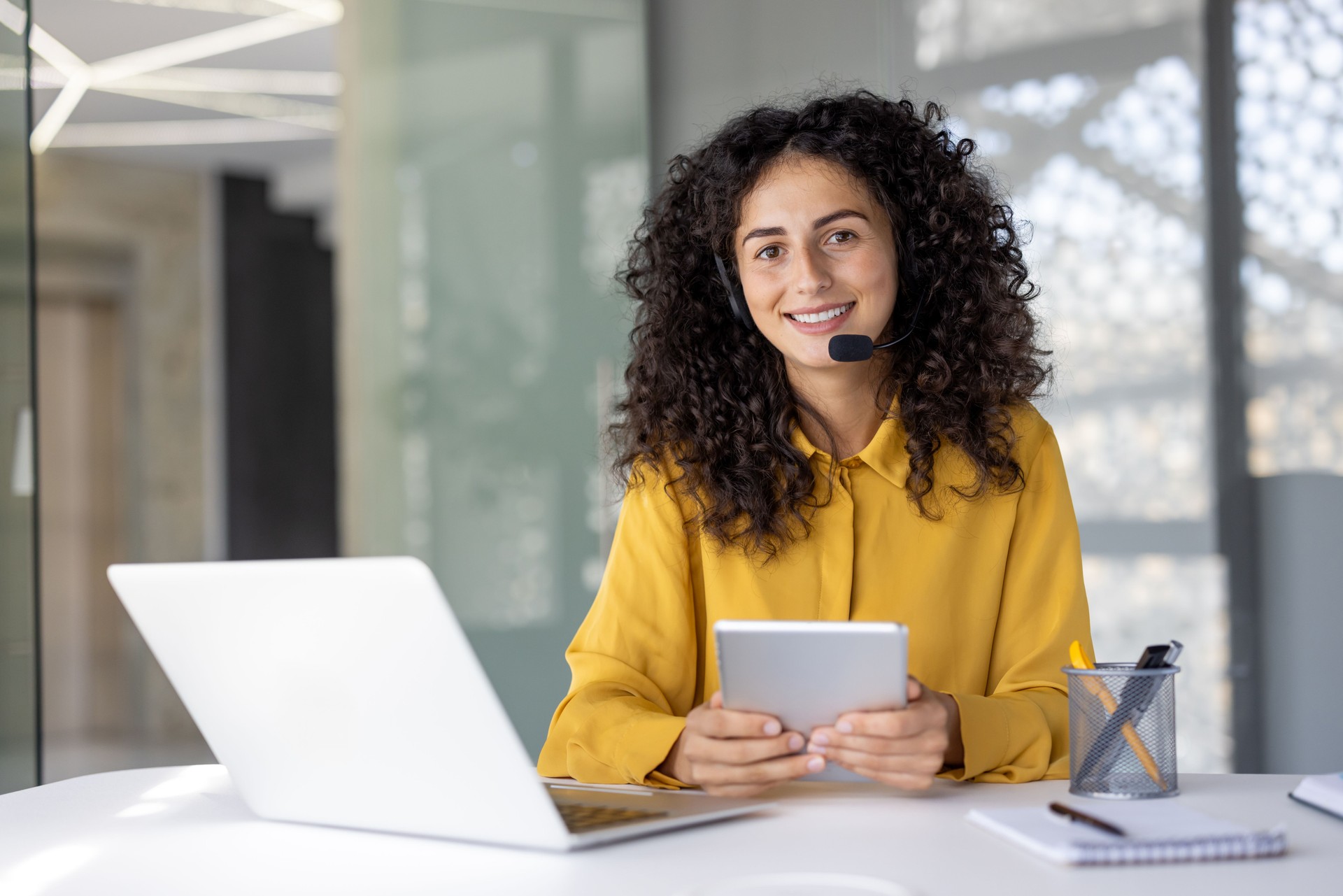 A woman with a headset smiles warmly while holding a tablet, ready to help customers with their inquiries.